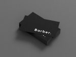 Printed black minimal personal barber business card held between two fingers in a barbershop setting, showing BARBER on the front face